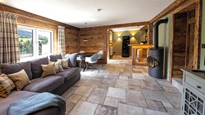 A cozy living room with a sofa, a dining table with chairs, and a stone floor. There is a fireplace with a chimney and a water dispenser in the background.