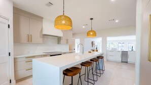 Una cocina moderna con armarios blancos, una isla grande con taburetes, iluminación colgante y vistas al área de comedor.