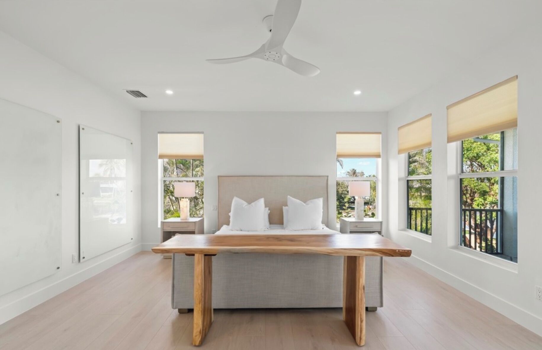 A modern bedroom with a large bed, a wooden table, and large windows with blinds.