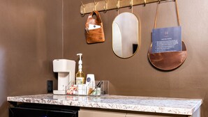 Un baño moderno con encimera de mármol, una cafetera, una botella de loción y un espejo.