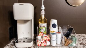 Una cafetera, una botella de sirope francés de vainilla Monin, y un organizador de plástico transparente con tazas y un cepillo de dientes sobre una encimera de mármol.