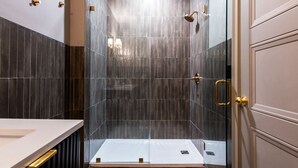 Un baño moderno con ducha envuelta en vidrio, paredes de azulejos oscuros y encimera blanca.
