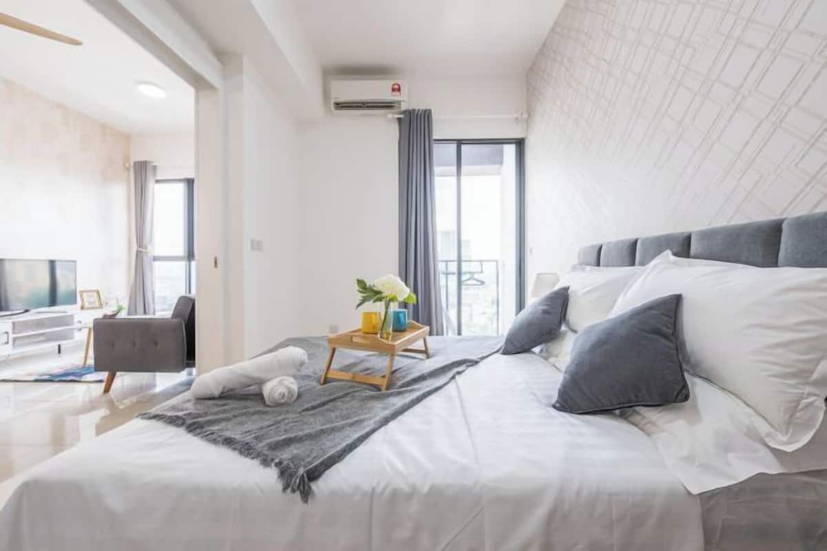 Ein modernes Schlafzimmer mit einem großen Bett, einem Nachttisch mit einer Vase und Tablett, einem grauen Sessel und Blick auf die Stadt durch ein großes Fenster.