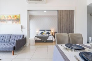 A modern hotel room with a bed, a sofa, a dining table with plates, and a painting on the wall. - Cozy Oasis near Ikea TRX Mall King Size Bed 100mb (Kuala Lumpur)