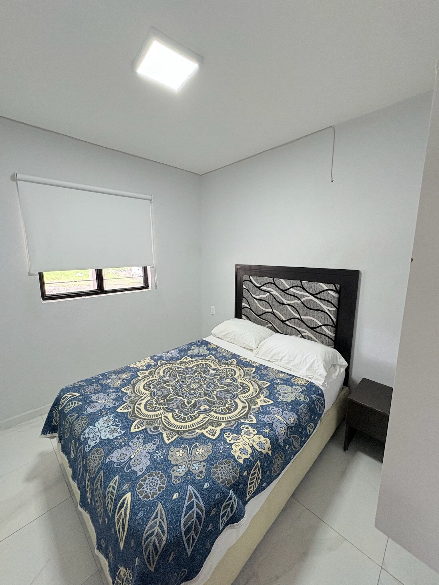 Une chambre à coucher avec un lit, une fenêtre avec des stores, une table de chevet et un luminaire au plafond.