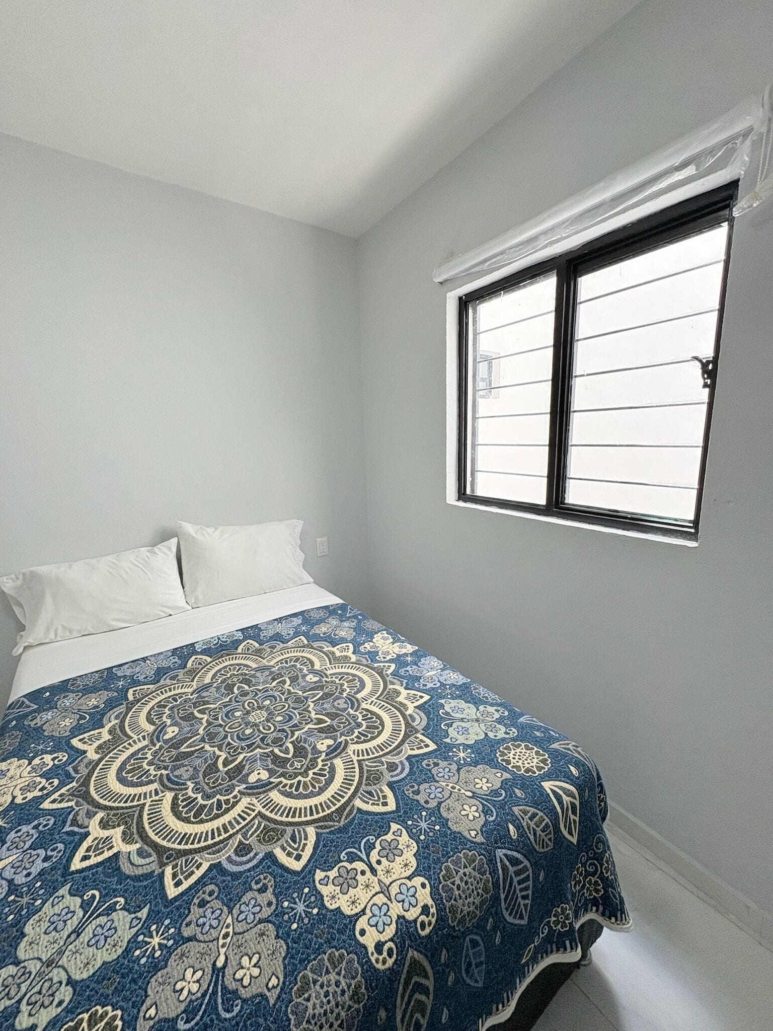 Une chambre à coucher avec un lit, des oreillers blancs, une couvre-lit à motifs bleus et dorés, et une fenêtre avec des stores.