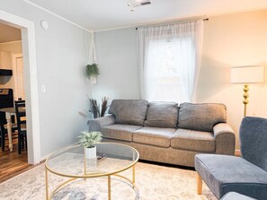 Un salon avec un canapé, une table basse ronde, une lampe et une fenêtre ornée de rideaux légers.