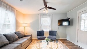 A living room with a sofa, two armchairs, a coffee table, a ceiling fan, a TV, and a window with curtains. - Cozy Beaufort Cottage - Near Parris Island & Downtown (Beaufort)