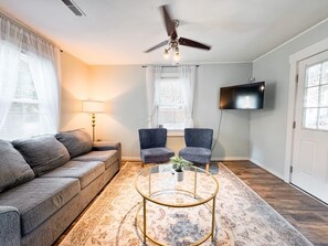 A living room with a sofa, two armchairs, a coffee table, a ceiling fan, a TV, and a window with curtains. - Cozy Beaufort Cottage - Near Parris Island & Downtown (Beaufort)