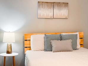 Une chambre à coucher avec une tête de lit en bois, une table de chevet ronde avec une lampe, et deux tableaux accrochés au mur.