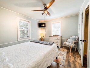 Une chambre à coucher comprenant un lit, un ventilateur de plafond, un téléviseur, une commode, une chaise et une fenêtre avec des stores.