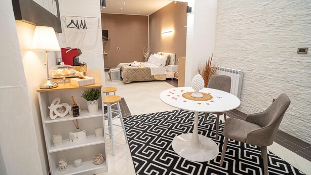 A modern hotel room with a bed, a round table with a decorative centerpiece, a small seating area with stools, and a white patterned rug.