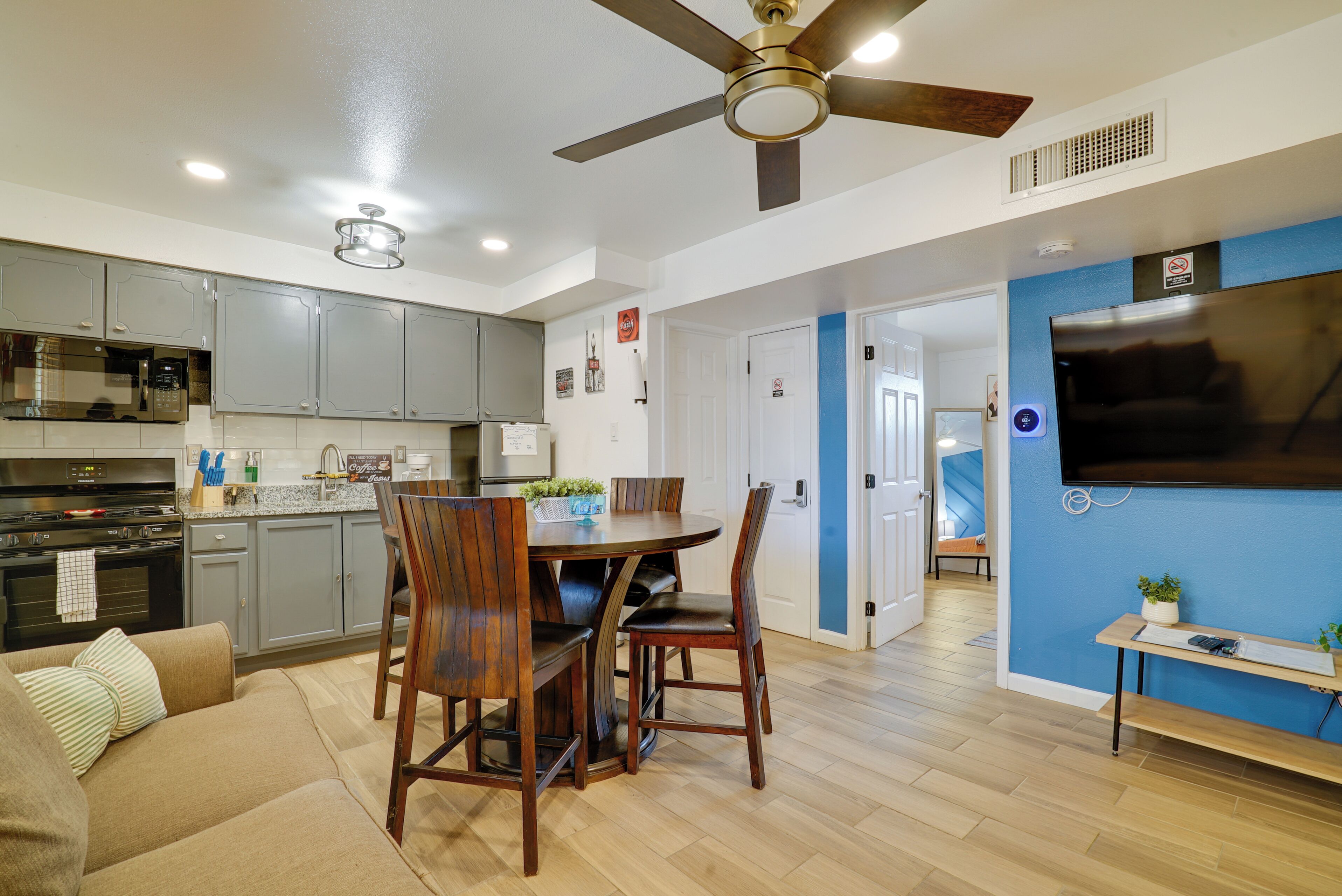 Una cocina moderna con gabinetes grises, una mesa de comedor con sillas, un ventilador de techo y un televisor montado en una pared azul.