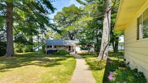 A house with a paved driveway leading to a front porch, surrounded by trees and a well-maintained lawn. - Lakefront Retreat w/ Pedal Boat & Dock in Kalkaska (Kalkaska)