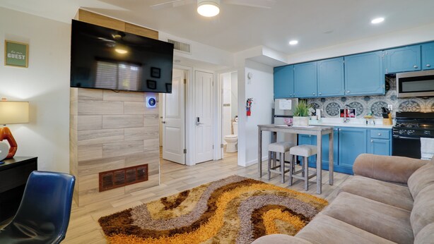 A modern living room with a sofa, a coffee table, and a TV. There is a kitchen area with blue cabinets and a microwave.