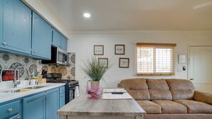 A kitchen with blue cabinets, a microwave, and a 'No Smoking' sign. There is a table with a plant and a remote control. A brown sofa is in the living area with a window and blinds.