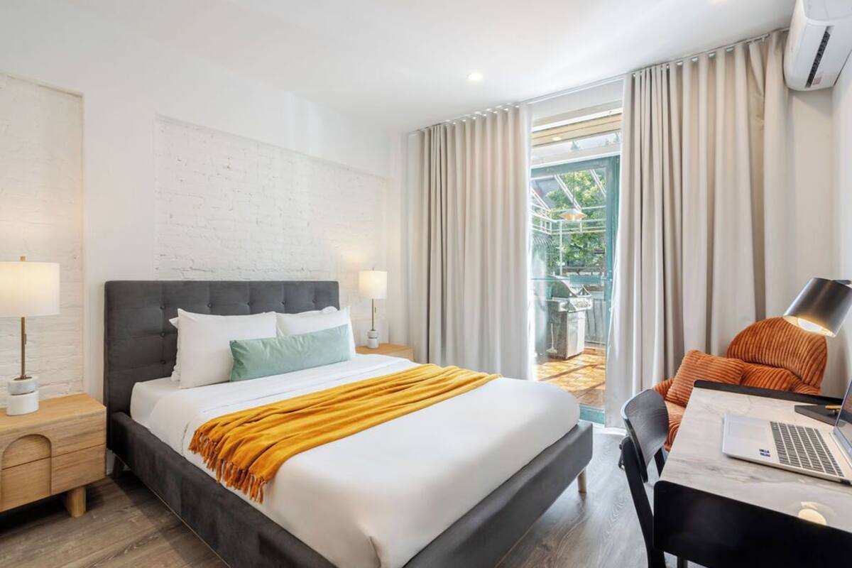 A modern hotel room with a large bed, a desk with a laptop, and a view of a garden through the window.