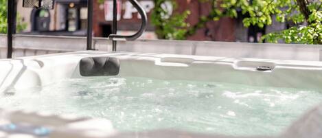 A hot tub with a metal faucet and a mesh cover, surrounded by greenery and a building in the background.