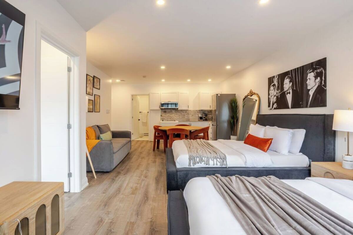 A modern hotel room with a large bed, a sofa, a dining area, and a kitchen in the background.