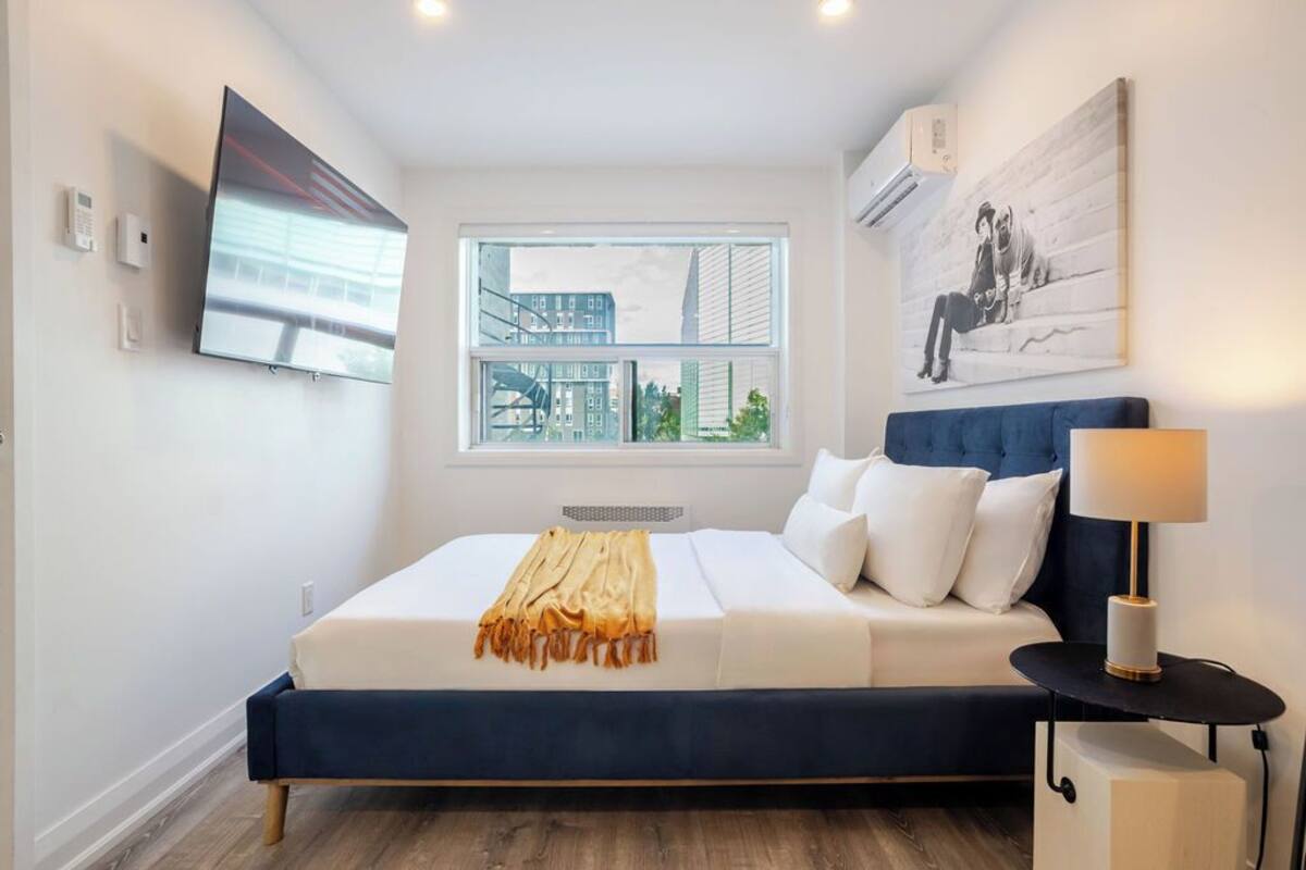 A modern bedroom with a large bed, a window with a city view, a mounted TV, and a wall-mounted air conditioner.