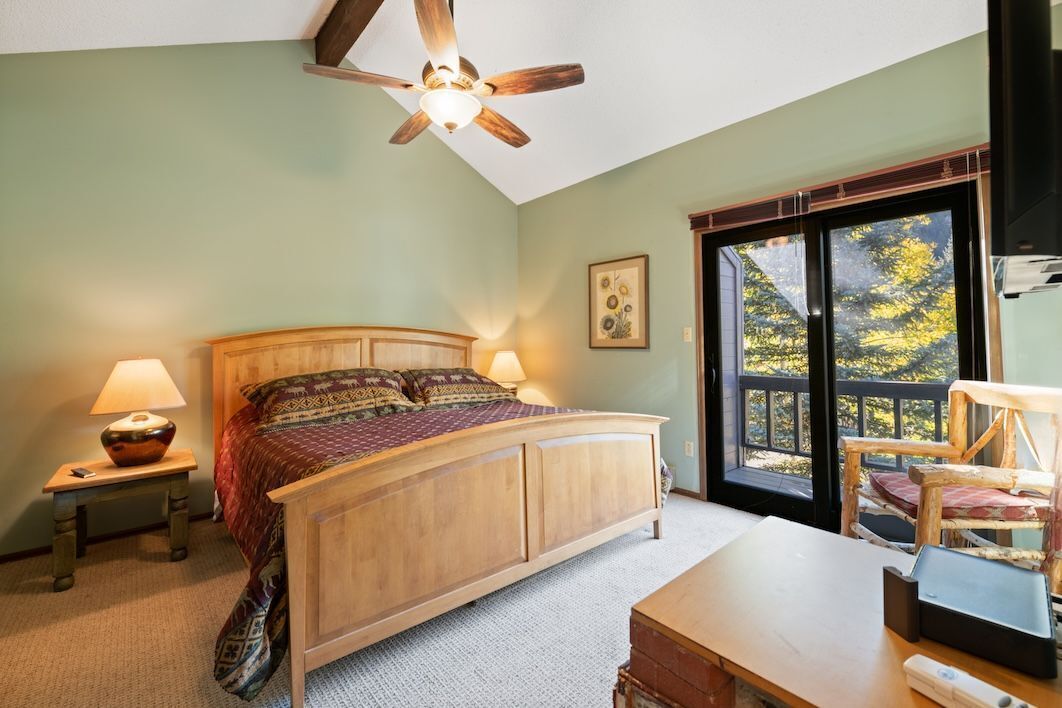 Une chambre avec un lit en bois, un ventilateur de plafond, une table de chevet avec une lampe, un bureau avec un ordinateur portable et une fenêtre donnant sur des arbres.