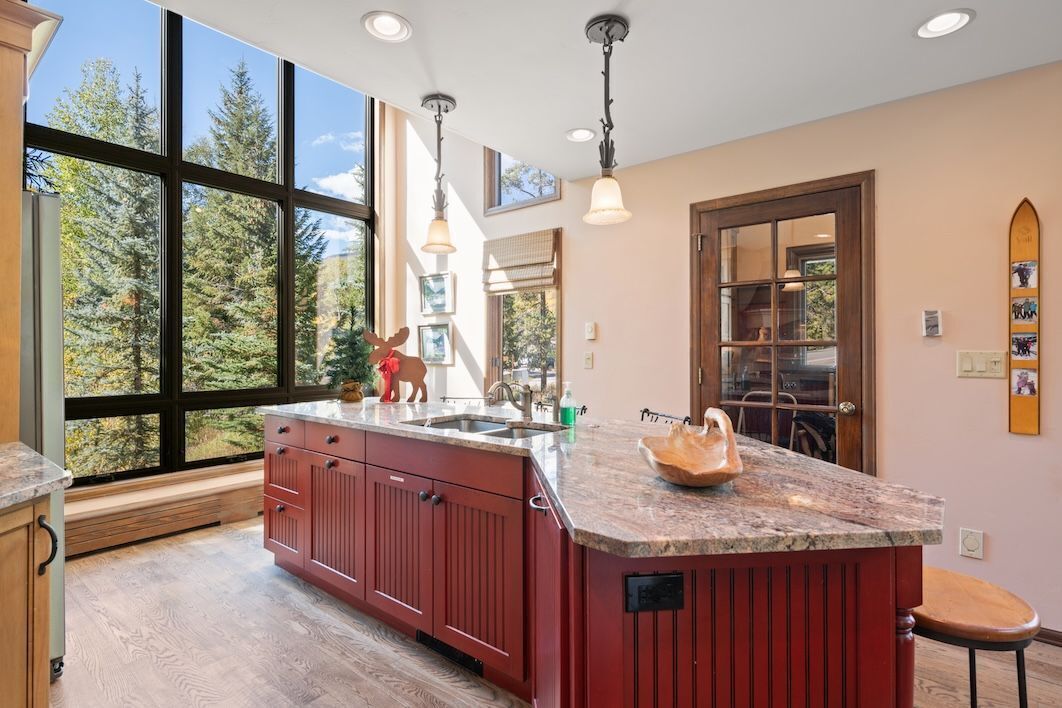 A kitchen with a large island, wooden cabinets, and a view of trees through large windows.