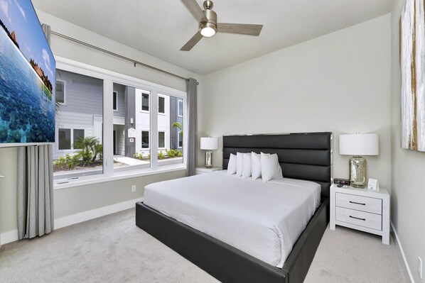 Un dormitorio con una cama grande, un ventilador de techo, una mesita de noche con lámpara y una ventana con vistas a edificios.