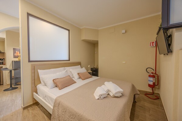 A neatly made bed with a beige bedspread and pillows, a fire extinguisher mounted on the wall, and a small desk with a chair and a plant. - Damas Home, apartment in St. Onofrio Square (Palermo)