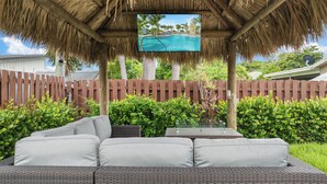 An outdoor seating area with a thatched roof, a pool view screen, and a wooden fence in the background.