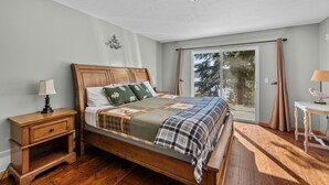 Un dormitorio con cama de madera, una mesita de noche, una lámpara, una ventana con cortinas y vistas a árboles.