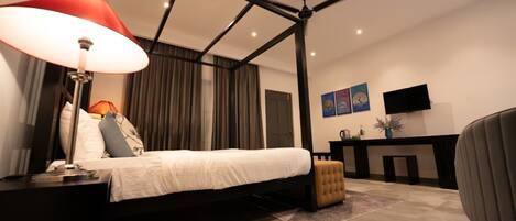A modern bedroom with a four-poster bed, a desk with a chair, a TV, and artwork on the wall.