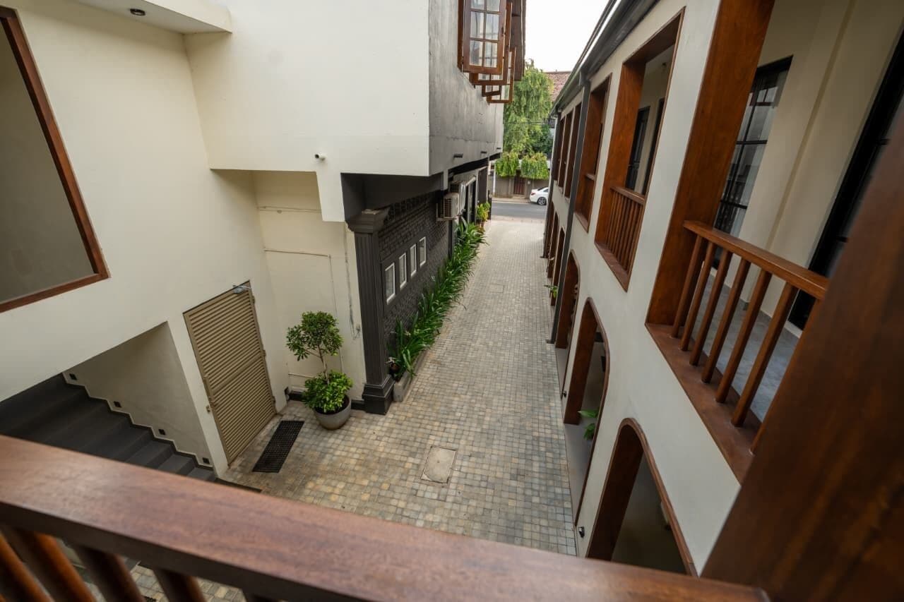 A narrow alleyway with a staircase on the left, a small garden area with plants, and buildings with wooden railings and windows on the right.