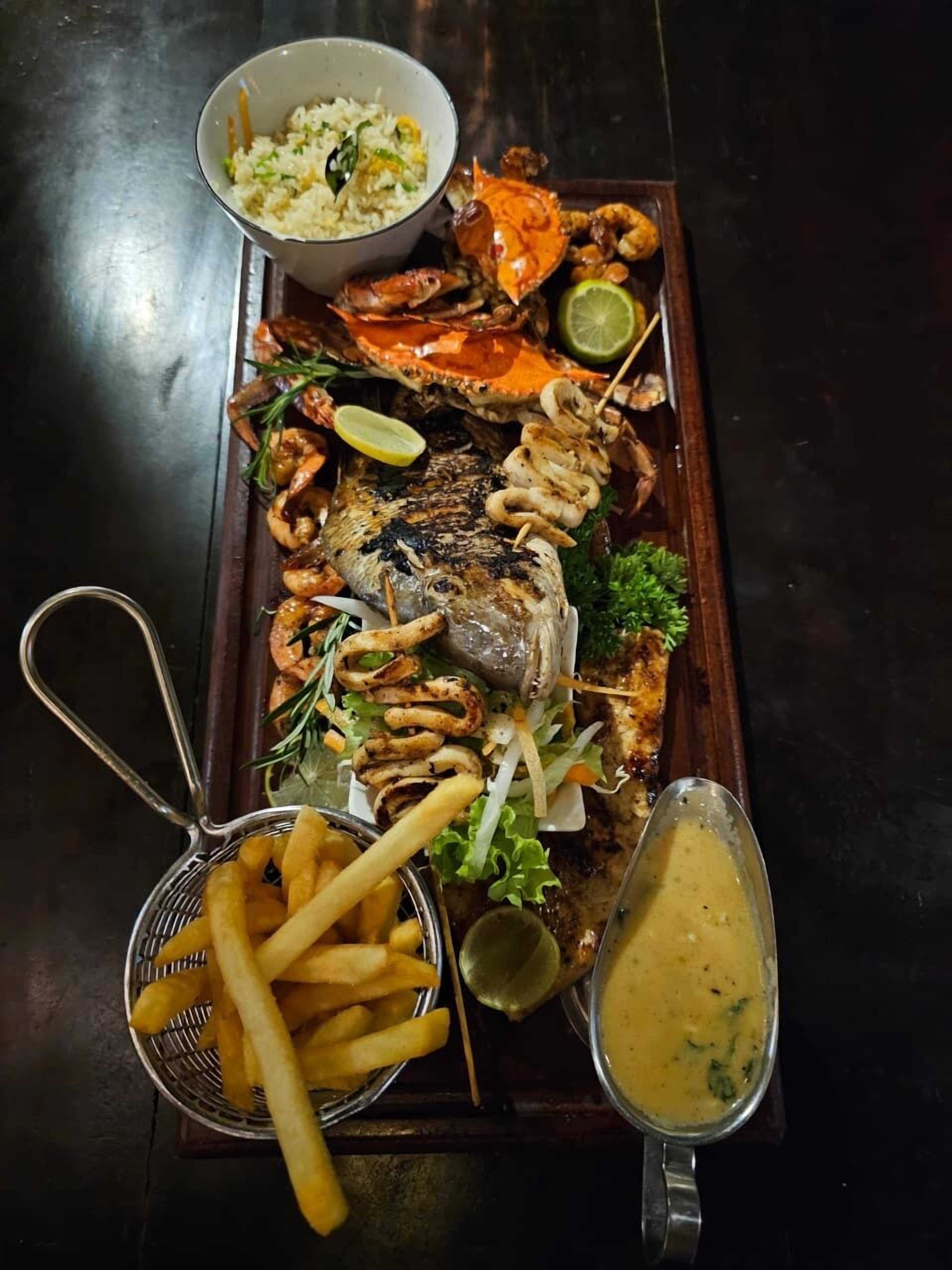 A seafood platter with grilled fish, shrimp, crab, and squid, accompanied by fries, coleslaw, and a dipping sauce.
