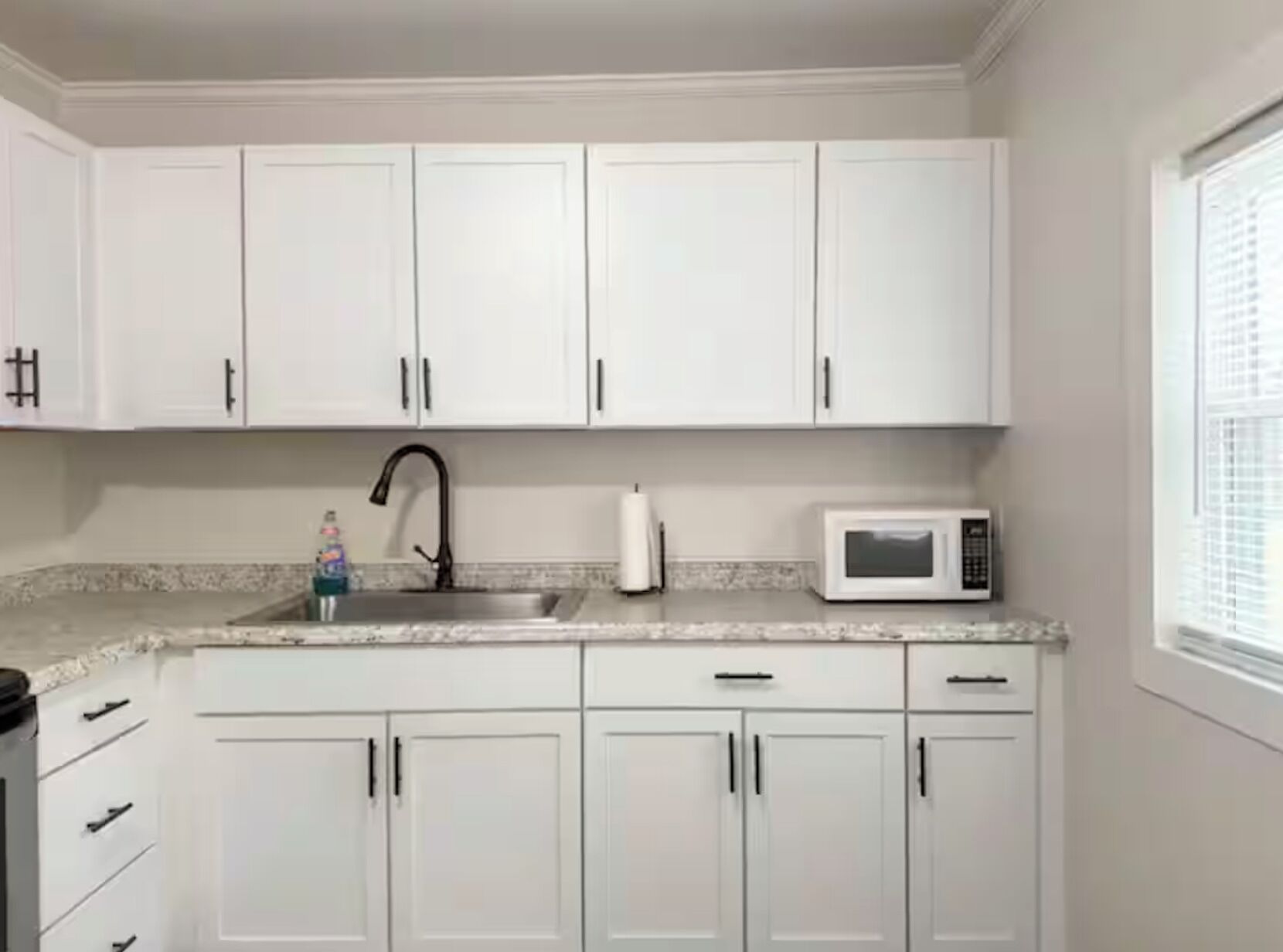 A kitchen with white cabinets, a stainless steel sink, a black faucet, a white microwave, and a window with blinds.