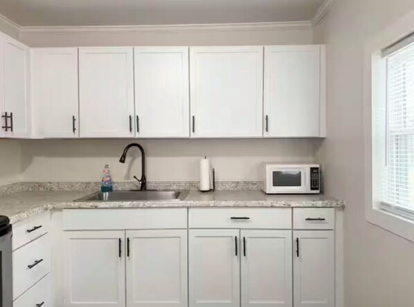 Une cuisine avec des meubles blancs, un évier en acier inoxydable, un robinet noir, un four à micro-ondes blanc et une fenêtre avec des stores.