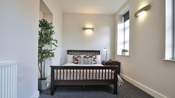 Quarto com cama, abajur, luz na parede, planta em vaso e janela.