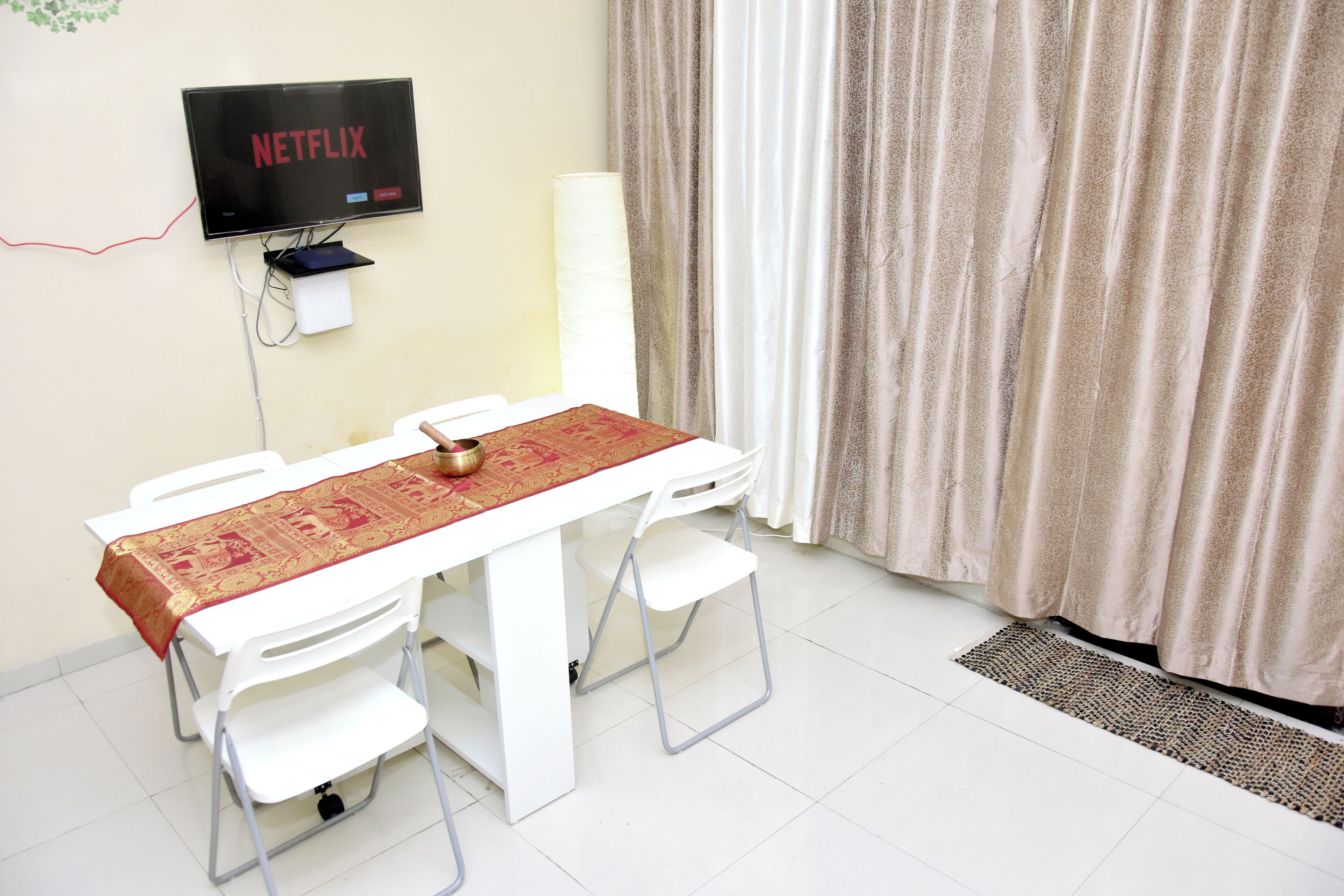 Une pièce avec une table blanche, des chaises blanches, un téléviseur fixé au mur et un rideau.