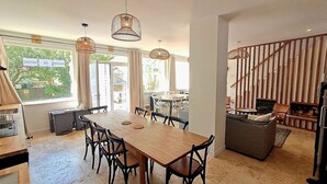 Uma sala de jantar ampla com mesa e cadeiras de madeira, janelas grandes e uma escada que dá para um mezanino.