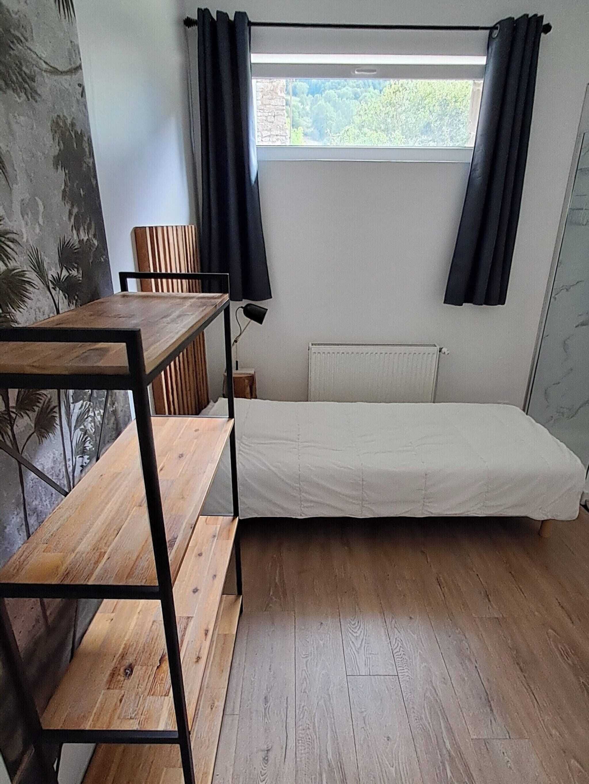 A small, single-room accommodation with a wooden floor, a simple bed, a table, and a chair.