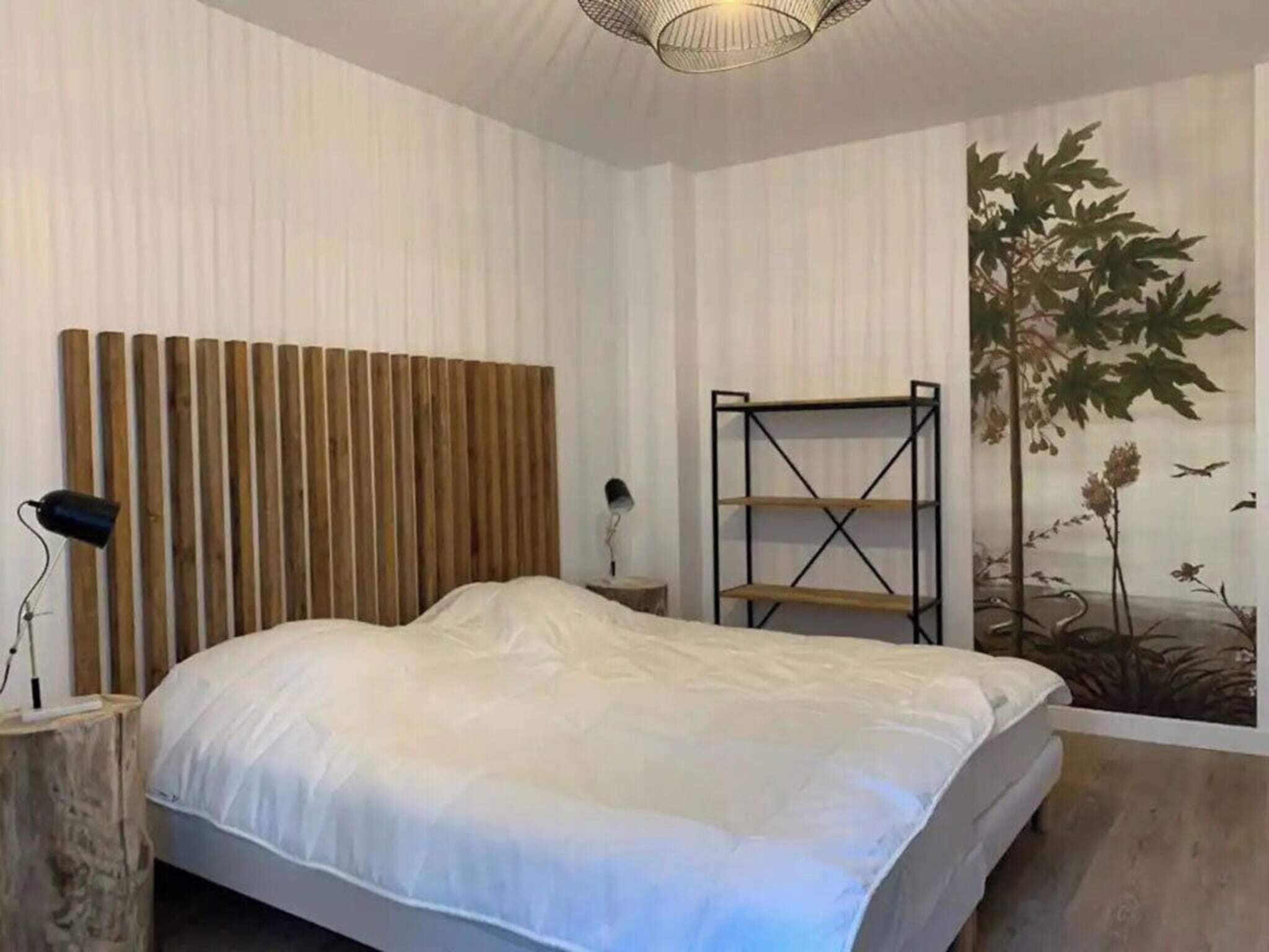 A bedroom with a bed, a wooden headboard, a nightstand, a lamp, a wall-mounted shelf, and a mural.