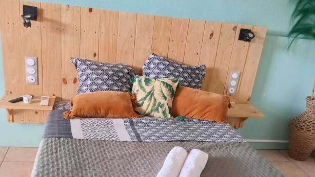 Ein Bett mit grauer, strukturierter Bettdecke, zwei aufgerollte Handtücher und ein Holz-Kopfteil mit gemusterten Kissen.