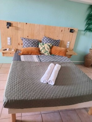 A bed with a grey textured bedspread, two rolled towels, and a wooden headboard with patterned pillows. - Le Swing - Studio (Saint-François)