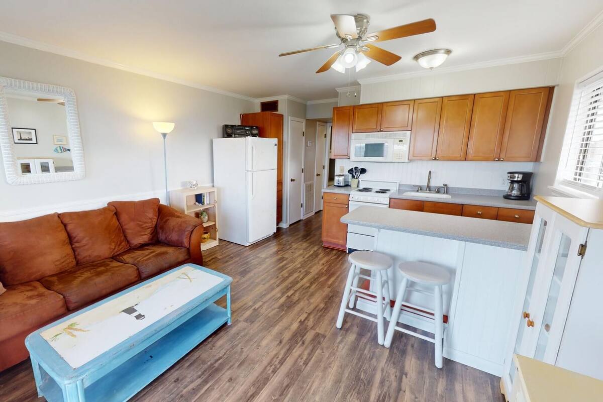 A compact living space with a brown sofa, a blue coffee table, a white refrigerator, a kitchen area with wooden cabinets, a microwave, and a ceiling fan.