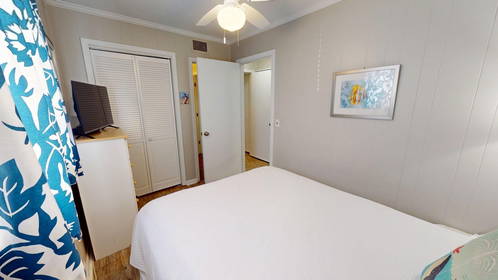 A bedroom with a bed, a wardrobe, a desk, a ceiling fan, and a door leading to another room.