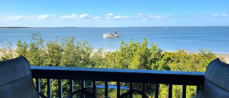 Un balcón con mesa y sillas, con vista a un mar tranquilo donde se distingue un barco a lo lejos y una playa al fondo.