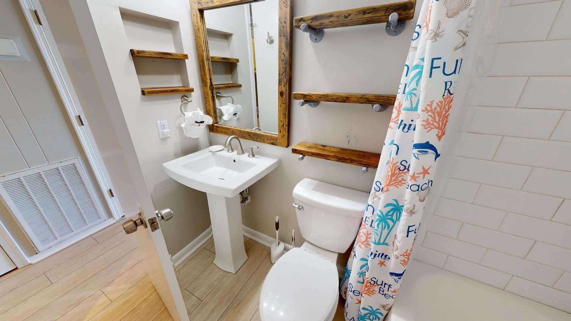 Un bagno con lavandino, water e tenda da doccia a tema spiaggia. Sopra il lavandino si trovano mensole in legno e uno specchio sulla parete.