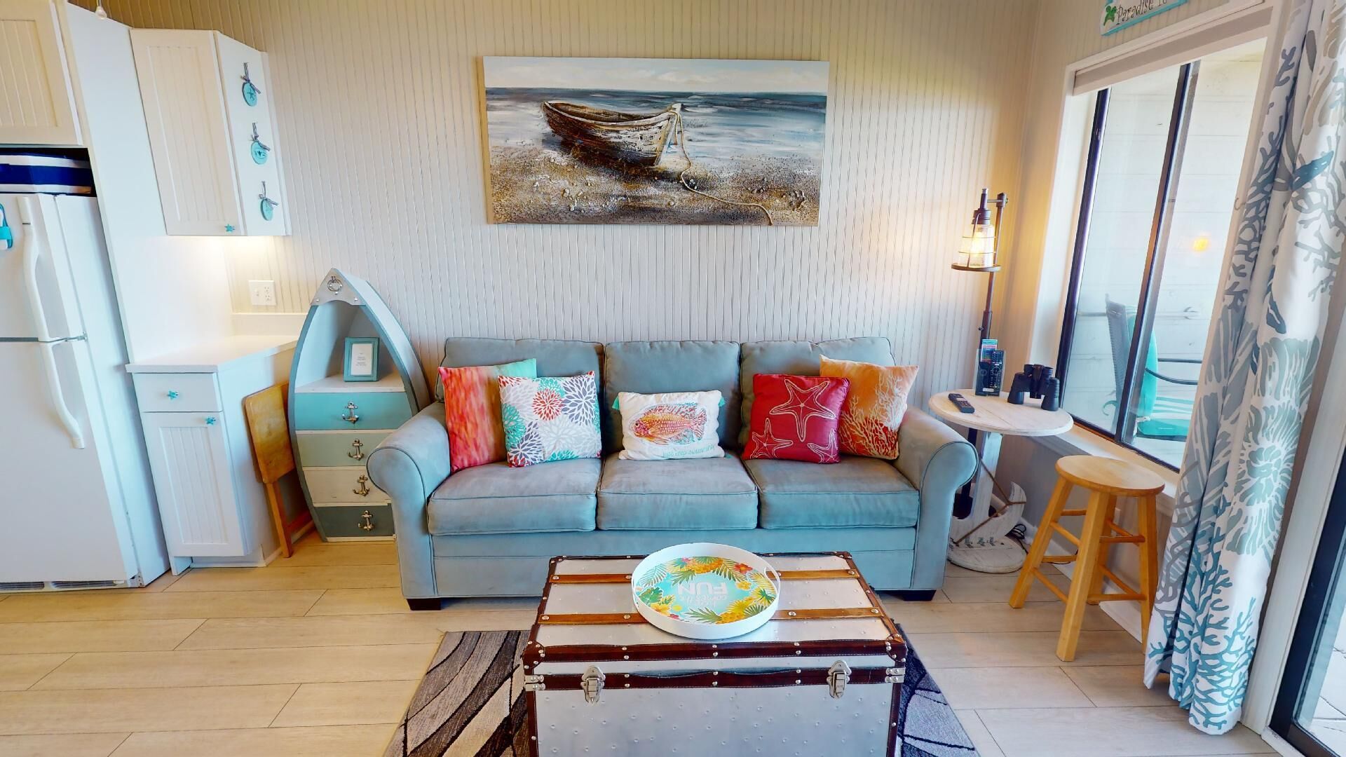 A cozy living room with a light blue sofa, colorful cushions, a vintage suitcase table, and a painting of a boat on the wall.