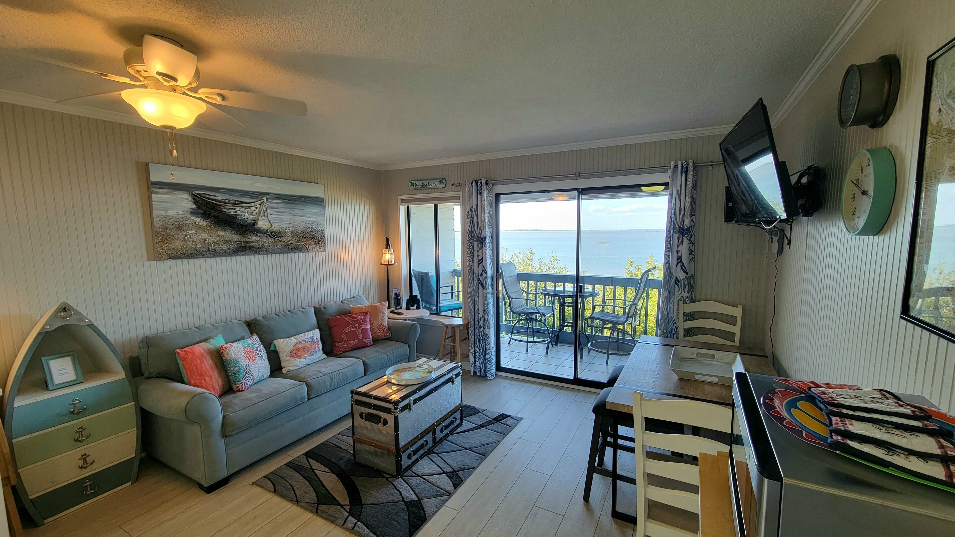 A living room with a sofa, coffee table, and a dining area with chairs and a table. There is a large window with a view of the sea.