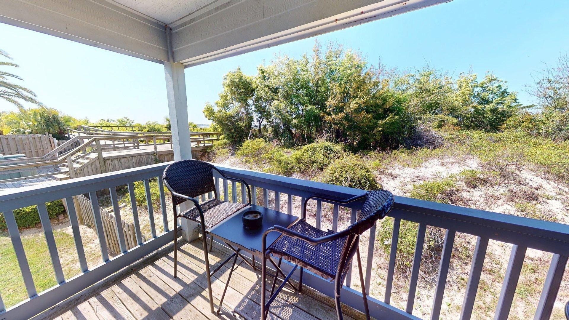 A covered outdoor area with a table and two chairs, overlooking a natural landscape with trees and shrubs.