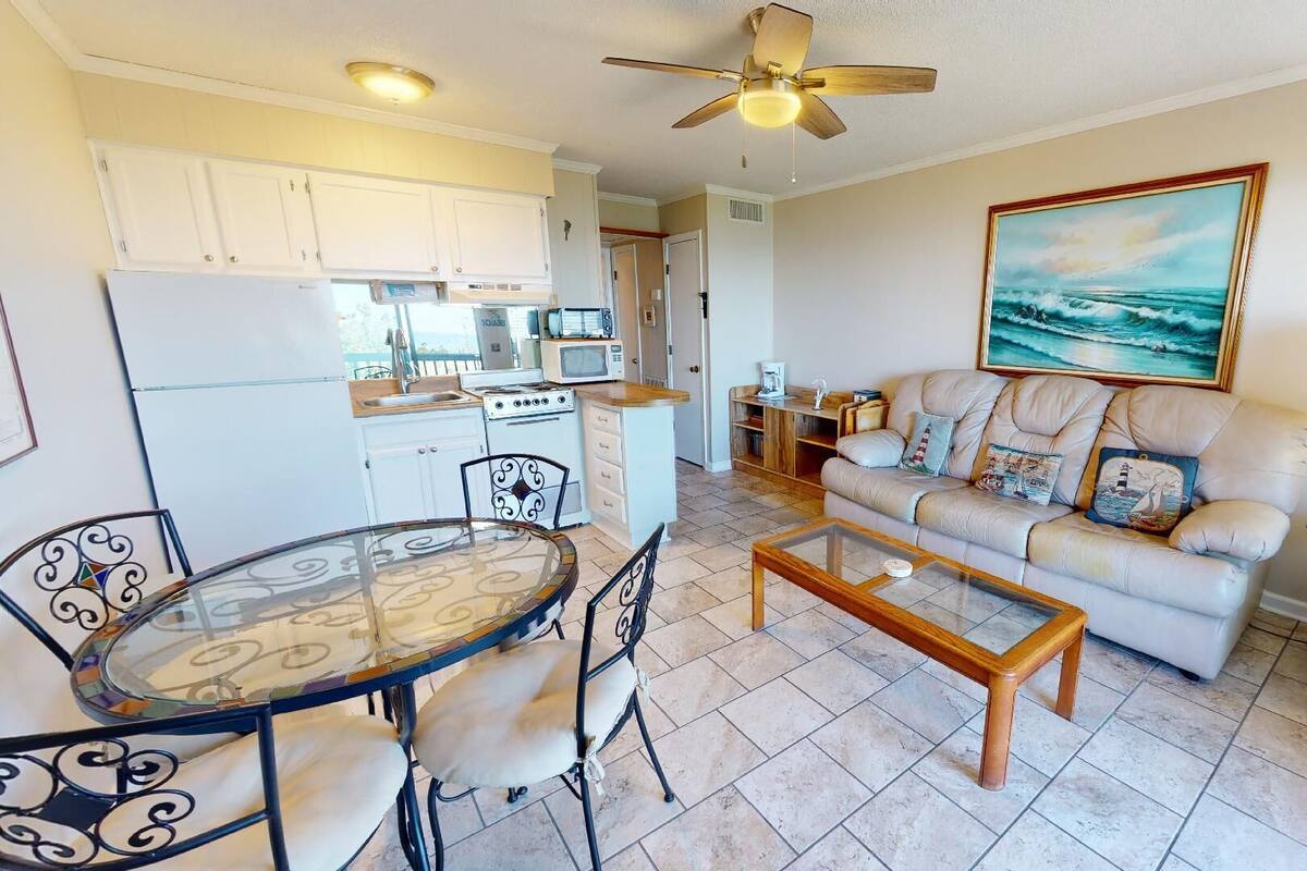 A compact living space with a kitchenette, a dining area, a sofa, and a painting of the sea.